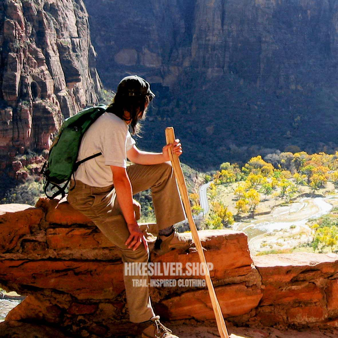 Hiker resting on canyon ledge with backpack and walking stick overlooking Zion valley and winding river below