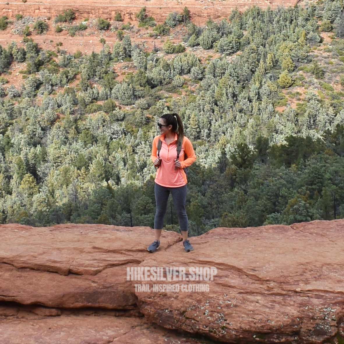 Woman hiker standing on red rock ledge in Sedona with forested canyon views and backpack gear-www.Hikesilver.shop